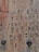 A close-up view of a wooden surface with various symbols and characters carved into it. The carvings appear to be of different styles, possibly representing a code or message. The wood has a weathered texture with horizontal grains and a few dark, rounded objects embedded in it.