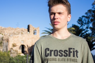A young person stands outdoors in sunlight, wearing a green CrossFit t-shirt. The background features ruins of a stone building with arched windows, along with greenery and trees in the scene.