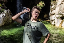 A person wearing a green CrossFit t-shirt holds a kettlebell over their shoulder near a rocky water area. The scene includes clear, green water, surrounding rocks, and a small waterfall in the background.