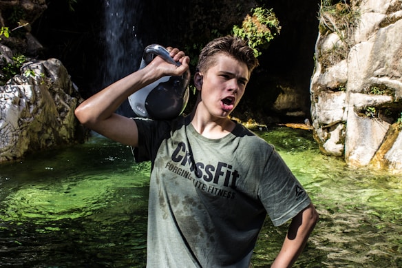 A person wearing a green CrossFit t-shirt holds a kettlebell over their shoulder near a rocky water area. The scene includes clear, green water, surrounding rocks, and a small waterfall in the background.