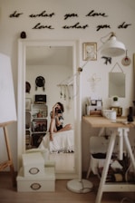A cozy room with minimalistic decor is visible. A woman is sitting on a bed in front of a mirror, taking a photo. The mirror reflects various decorative items such as wall art, plants, and shelves with personal belongings. A motivational quote is displayed on the wall above the mirror, reading 'do what you love, love what you do'. The space includes a desk with art supplies and a lamp, creating a creative and personal atmosphere.