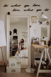 A cozy room with minimalistic decor is visible. A woman is sitting on a bed in front of a mirror, taking a photo. The mirror reflects various decorative items such as wall art, plants, and shelves with personal belongings. A motivational quote is displayed on the wall above the mirror, reading 'do what you love, love what you do'. The space includes a desk with art supplies and a lamp, creating a creative and personal atmosphere.