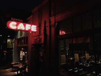 A dimly lit street scene featuring a cafe with a neon sign reading 'CAFE'. The sign is glowing brightly in contrast to the dark surroundings. Inside the cafe, visible through the large front window, chairs are stacked on tables indicating that the cafe is likely closed. The building exterior has an urban ambiance with decorative elements.