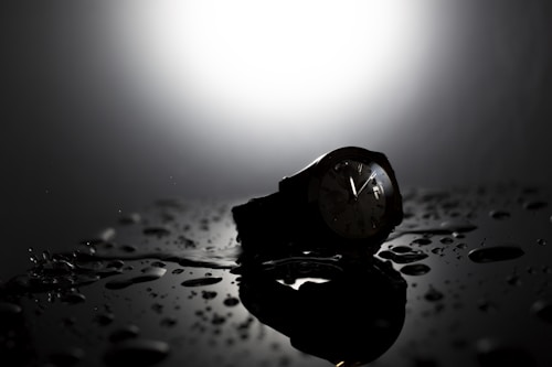 A dark and moody close-up of a wristwatch resting on a reflective surface with water droplets scattered around. The watch face is illuminated by a bright, focused light from above, creating a dramatic silhouette effect and enhancing the contrast between light and shadow.