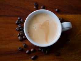 A cup of creamy espresso sits on a wooden surface, accompanied by scattered coffee beans. The rich texture of the coffee foam is highlighted against the dark, polished wood.