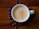 A cup of creamy espresso sits on a wooden surface, accompanied by scattered coffee beans. The rich texture of the coffee foam is highlighted against the dark, polished wood.