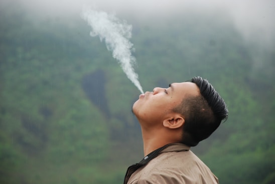 A person is outdoors exhaling a stream of smoke or vapor upwards, set against a blurred, verdant landscape background. The individual appears calm and contemplative.