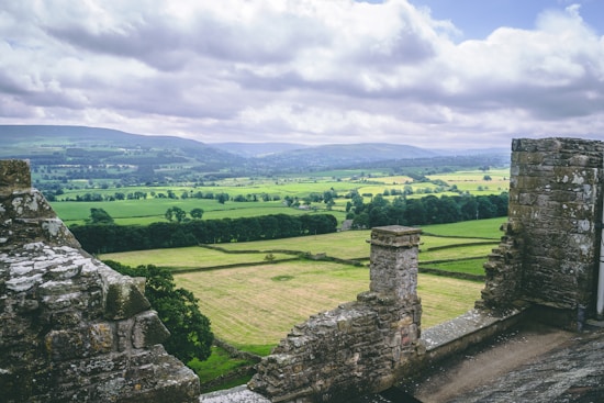 Ruins of an ancient stone structure overlook a vast and lush green landscape. Rolling hills and a patchwork of fields stretch into the distance beneath a cloudy sky.