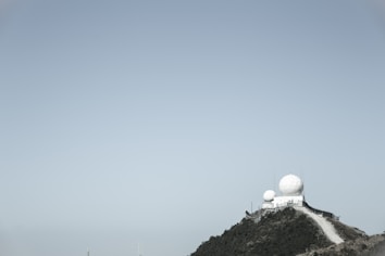 A hilltop installation with large white radar domes surrounded by a security fence. The sky is clear, and the landscape is mostly barren with some shrubbery.