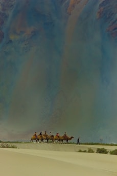 A group of people riding camels traverse across a vast desert landscape. The sand is marked by gentle dunes, and the sky above appears vast and expansive, possibly indicating a time of day when the sun is low. The scene conveys a sense of adventure and exploration.