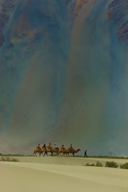 A group of people riding camels traverse across a vast desert landscape. The sand is marked by gentle dunes, and the sky above appears vast and expansive, possibly indicating a time of day when the sun is low. The scene conveys a sense of adventure and exploration.
