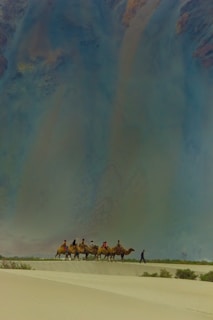 A group of people riding camels traverse across a vast desert landscape. The sand is marked by gentle dunes, and the sky above appears vast and expansive, possibly indicating a time of day when the sun is low. The scene conveys a sense of adventure and exploration.