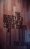 Large black text on a wooden floor spells out the message about the importance of soil in coffee quality. The text is bold and capitalized, creating a strong visual contrast against the wood grain background.