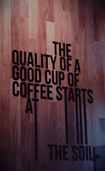 Large black text on a wooden floor spells out the message about the importance of soil in coffee quality. The text is bold and capitalized, creating a strong visual contrast against the wood grain background.