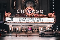 An illuminated theater marquee displays upcoming shows, with bright lights and a classic design. The marquee highlights events for Dream Theater on November 3 and Peppa Pig Live on November 4. A pickup truck is parked on the wet street in front of the theater, reflecting the evening ambiance.