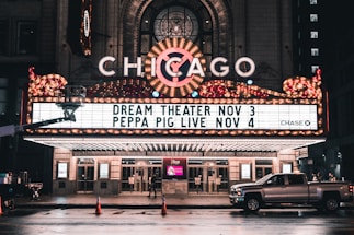 An illuminated theater marquee displays upcoming shows, with bright lights and a classic design. The marquee highlights events for Dream Theater on November 3 and Peppa Pig Live on November 4. A pickup truck is parked on the wet street in front of the theater, reflecting the evening ambiance.