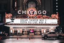 An illuminated theater marquee displays upcoming shows, with bright lights and a classic design. The marquee highlights events for Dream Theater on November 3 and Peppa Pig Live on November 4. A pickup truck is parked on the wet street in front of the theater, reflecting the evening ambiance.