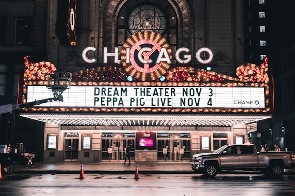 An illuminated theater marquee displays upcoming shows, with bright lights and a classic design. The marquee highlights events for Dream Theater on November 3 and Peppa Pig Live on November 4. A pickup truck is parked on the wet street in front of the theater, reflecting the evening ambiance.
