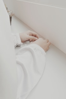 Hands placed on a white surface, wearing long white sleeves, in a minimalist setting with a small plant partially visible on one side.