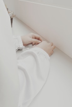 Hands placed on a white surface, wearing long white sleeves, in a minimalist setting with a small plant partially visible on one side.