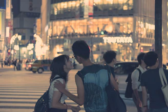A stylish couple sharing a quiet, intimate moment under soft purple and red city lights at night.
