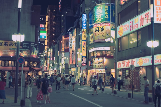A bustling urban street scene at night, with many neon signs in Japanese lighting up the surroundings. People walk along the street, some in groups and others alone, creating a vibrant and lively atmosphere.