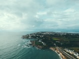A coastal landscape features a rocky shoreline with ocean waves meeting the land. A cluster of houses stretches along the coastline, with more residential areas visible in the distance. The scenery includes lush green fields and patches of trees, with a road extending along the edge of the land. The sky is overcast with a mix of clouds, creating a moody yet serene atmosphere.