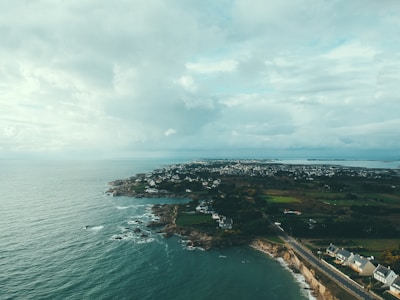 A coastal landscape features a rocky shoreline with ocean waves meeting the land. A cluster of houses stretches along the coastline, with more residential areas visible in the distance. The scenery includes lush green fields and patches of trees, with a road extending along the edge of the land. The sky is overcast with a mix of clouds, creating a moody yet serene atmosphere.