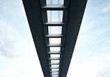 A symmetrical perspective view of the underside of a modern bridge, featuring a series of evenly spaced beams and support structures. The sky is visible through the gaps between the beams, adding a contrast between the solid structure and the open sky. The image conveys a sense of architectural elegance and engineering precision.