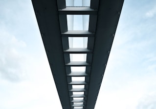A symmetrical perspective view of the underside of a modern bridge, featuring a series of evenly spaced beams and support structures. The sky is visible through the gaps between the beams, adding a contrast between the solid structure and the open sky. The image conveys a sense of architectural elegance and engineering precision.