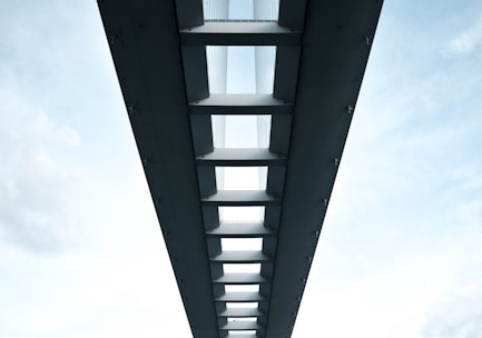 A symmetrical perspective view of the underside of a modern bridge, featuring a series of evenly spaced beams and support structures. The sky is visible through the gaps between the beams, adding a contrast between the solid structure and the open sky. The image conveys a sense of architectural elegance and engineering precision.