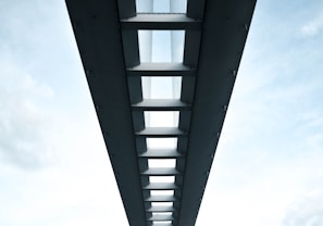 A symmetrical perspective view of the underside of a modern bridge, featuring a series of evenly spaced beams and support structures. The sky is visible through the gaps between the beams, adding a contrast between the solid structure and the open sky. The image conveys a sense of architectural elegance and engineering precision.
