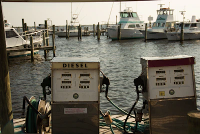 Two vintage diesel fuel pumps are situated on a wooden dock with a backdrop of several docked boats in a marina. The water is lightly rippling under a soft, warm light.