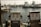 Two vintage diesel fuel pumps are situated on a wooden dock with a backdrop of several docked boats in a marina. The water is lightly rippling under a soft, warm light.