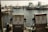 Two vintage diesel fuel pumps are situated on a wooden dock with a backdrop of several docked boats in a marina. The water is lightly rippling under a soft, warm light.