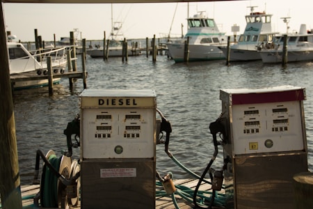 Two vintage diesel fuel pumps are situated on a wooden dock with a backdrop of several docked boats in a marina. The water is lightly rippling under a soft, warm light.