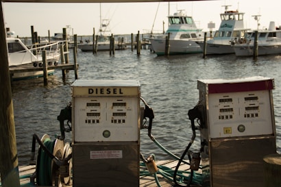 Two vintage diesel fuel pumps are situated on a wooden dock with a backdrop of several docked boats in a marina. The water is lightly rippling under a soft, warm light.