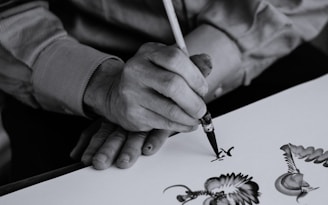 A person is skillfully painting intricate designs with a fine brush on a white surface, demonstrating precision and artistic flair. The focus is on the detailed artwork and the careful hand movements of the artist.