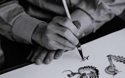 A person is skillfully painting intricate designs with a fine brush on a white surface, demonstrating precision and artistic flair. The focus is on the detailed artwork and the careful hand movements of the artist.