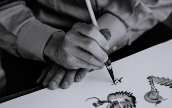A person is skillfully painting intricate designs with a fine brush on a white surface, demonstrating precision and artistic flair. The focus is on the detailed artwork and the careful hand movements of the artist.
