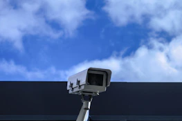 Technician installing a security camera on a large construction framework against a clear blue sky.
