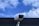 A security camera is mounted on a pole against a backdrop of a blue sky with scattered white clouds. The camera is positioned at an angle, suggesting it is set up for surveillance. The clear sky provides a contrasting background to the metallic structure of the camera.