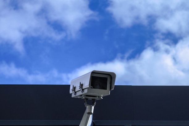 A technician installing a security camera on a business storefront under a clear sky.