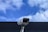 A security camera is mounted on a pole against a backdrop of a blue sky with scattered white clouds. The camera is positioned at an angle, suggesting it is set up for surveillance. The clear sky provides a contrasting background to the metallic structure of the camera.