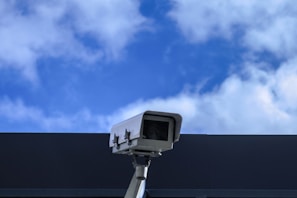 A security camera is mounted on a pole against a backdrop of a blue sky with scattered white clouds. The camera is positioned at an angle, suggesting it is set up for surveillance. The clear sky provides a contrasting background to the metallic structure of the camera.