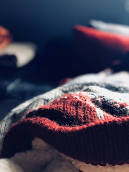 A close-up of a knitted fabric with a warm red hue and intricate patterns illuminated by soft light. The background contains blurred dark and light areas, suggesting a cozy interior setting.