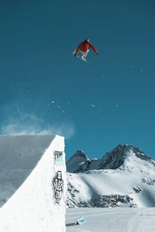 A snowscoot rider launching off a snowy mountain ramp with a dramatic alpine backdrop.