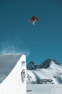 A snowboarder is captured in mid-air against a clear blue sky, performing a high jump off a snow ramp. Snow-covered mountains loom in the background, adding a sense of scale and adventure. The ramp shows branding and logo stickers, indicating possible sponsorship or event participation.