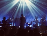 A band is performing on stage with atmospheric lighting. The musicians are playing various instruments including drums and keyboards. Beams of light create a dramatic effect, and the audience is visible as silhouettes in the foreground.