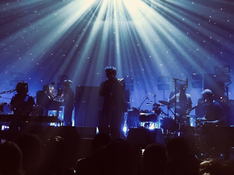 A band is performing on stage with atmospheric lighting. The musicians are playing various instruments including drums and keyboards. Beams of light create a dramatic effect, and the audience is visible as silhouettes in the foreground.